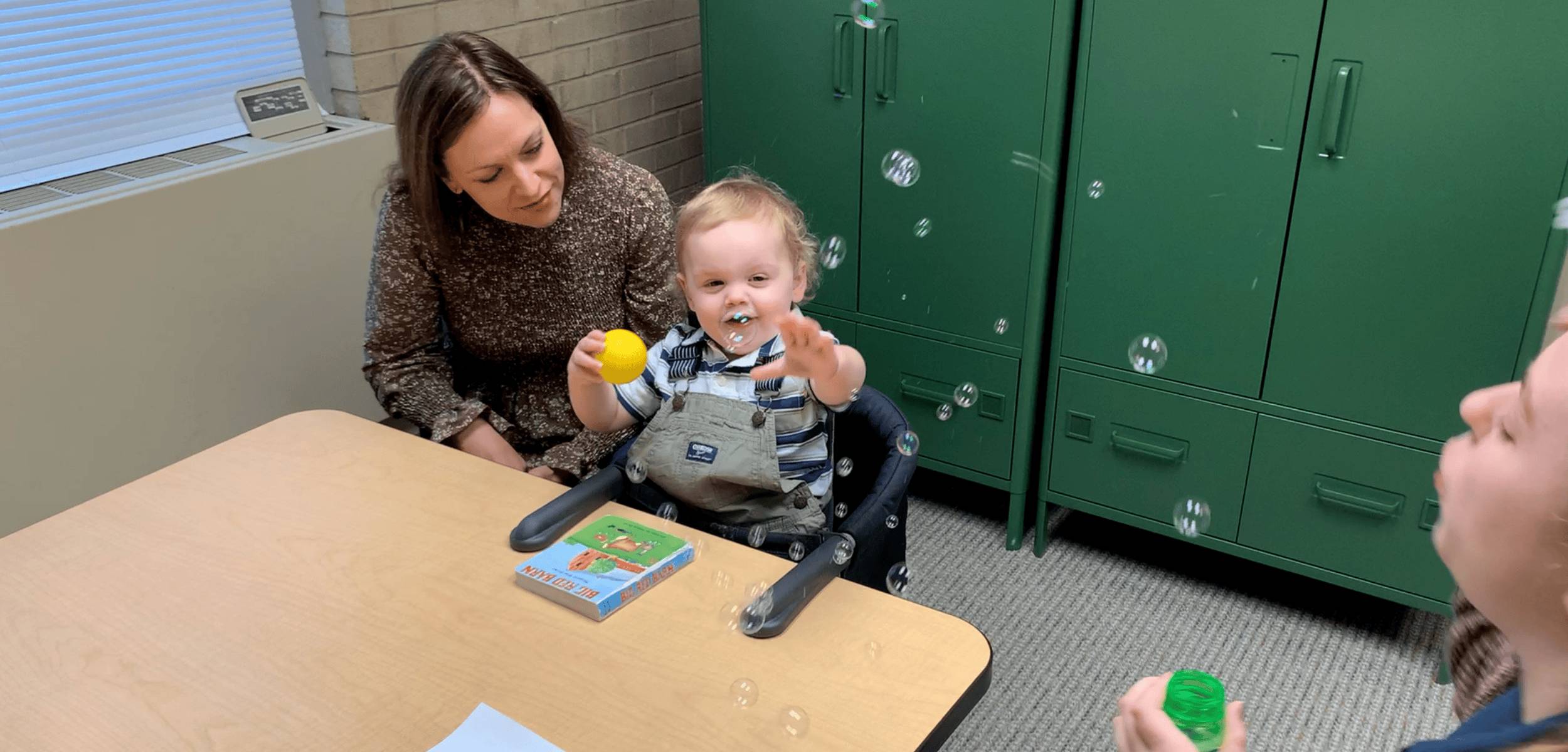 Toddler reaching for bubbles during a developmental play activity while a clinician observes.