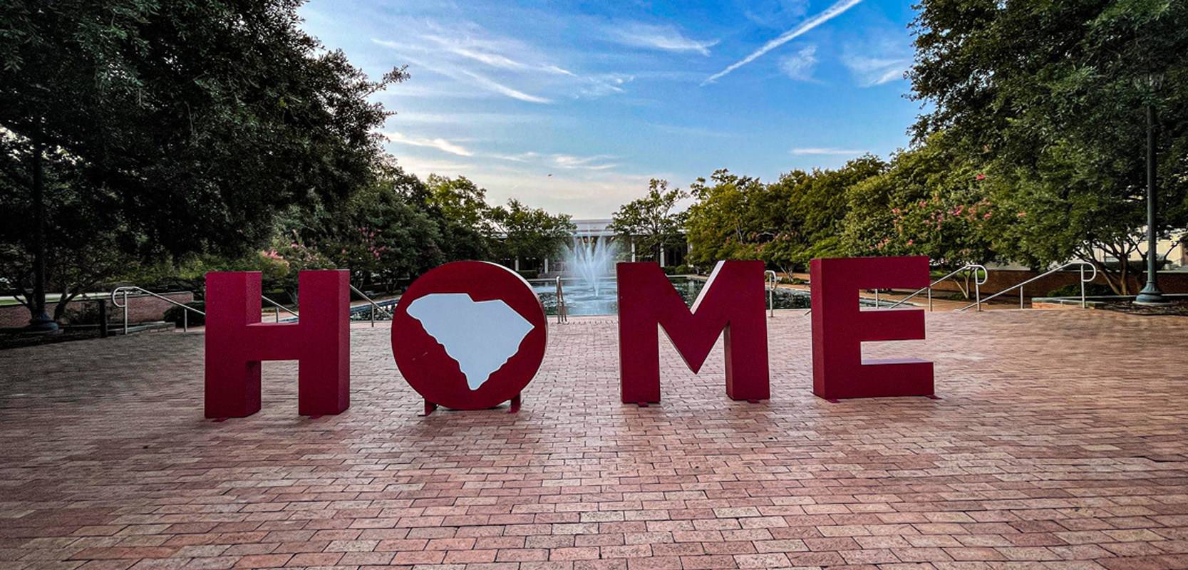 Large garnet letters spelling “HOME” with the South Carolina state outline replacing the “O,” displayed in front of the Thomas Cooper Library fountain on the University of South Carolina campus at sunset.