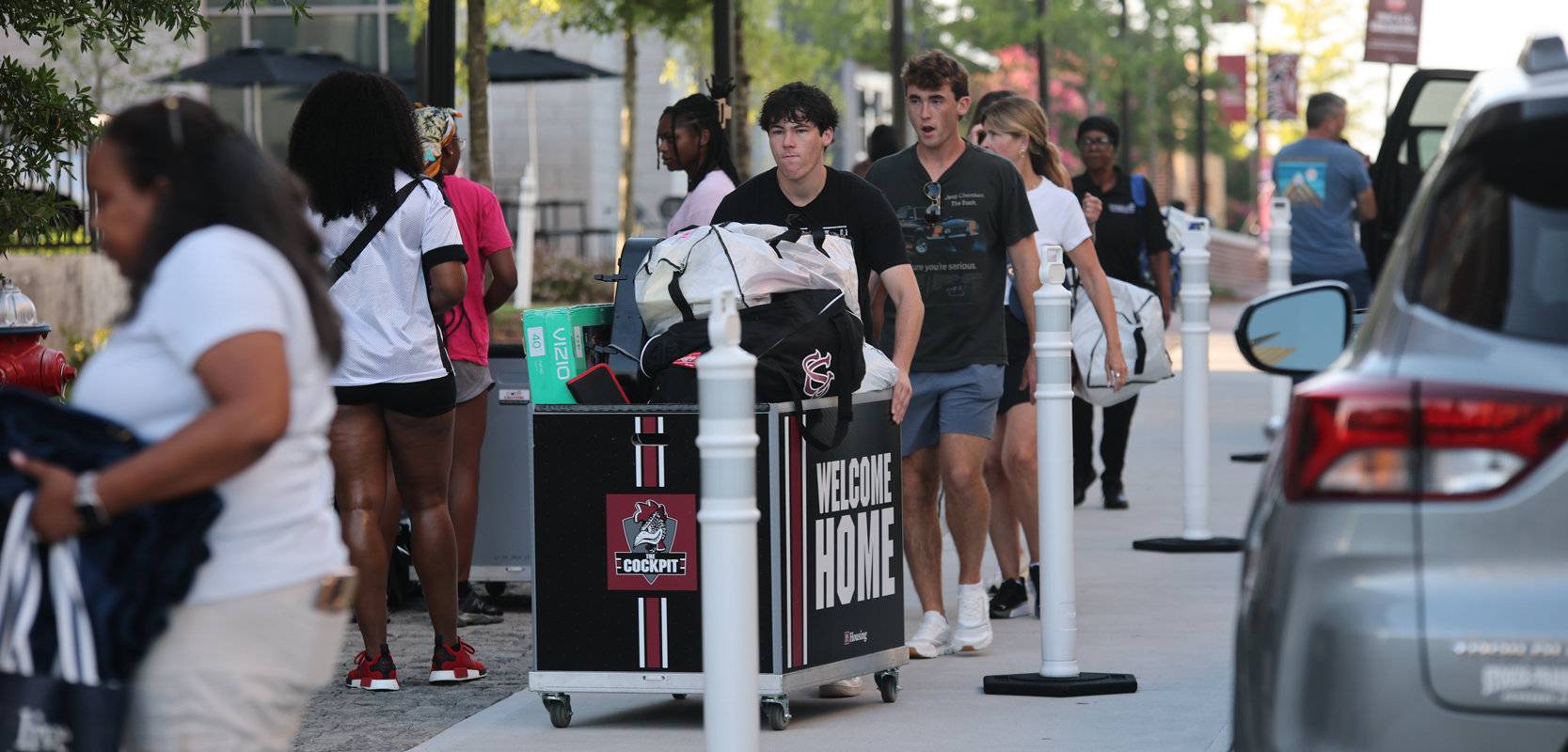 Students and family members move belongings in large “Welcome Home” carts along a campus sidewalk during University of South Carolina move-in day, with cars parked nearby for unloading.