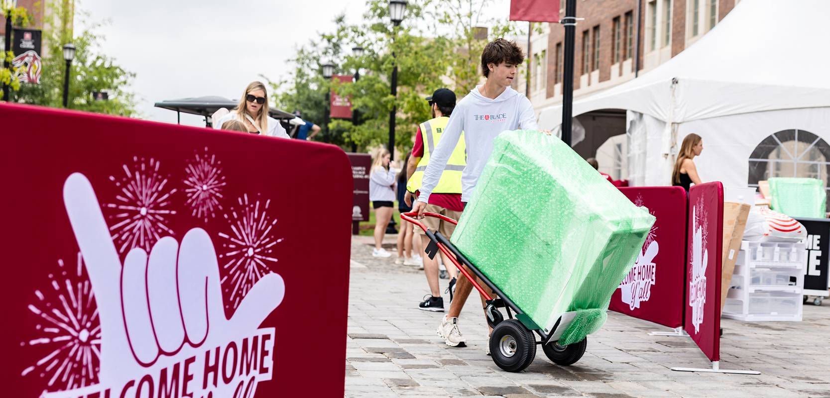 University of South Carolina move-in day at Campus Village, with a student pushing a dolly loaded with green-wrapped items past garnet ‘Welcome Home Y’all’ signs, volunteers, and tents in the background.