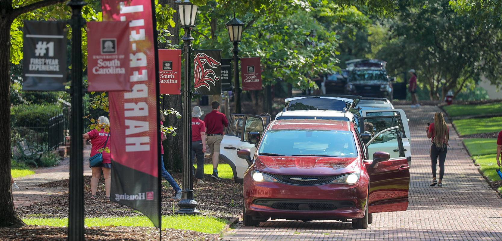 Families and students participate in move-in day at the University of South Carolina, unloading vehicles along a tree-lined brick road with campus banners and volunteers in garnet shirts offering assistance.