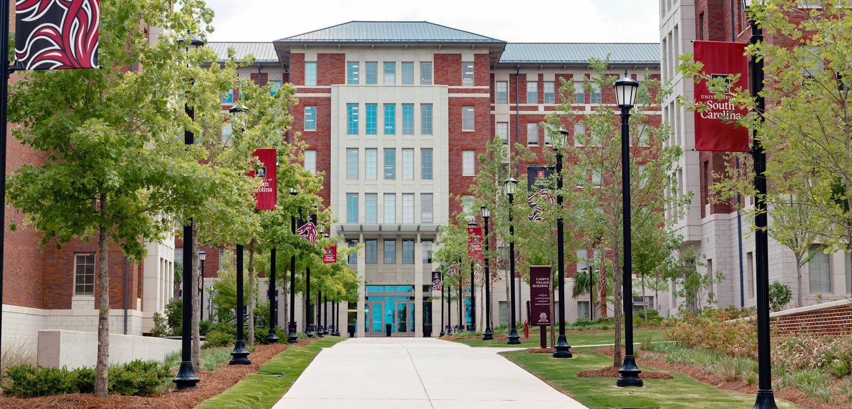 A walk way lined with trees and South Carolina banners on light poles leads you to a six story brick residence hall, Campus Village 4