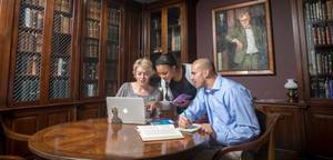 Three people collaborate around a laptop at a table in a library or study room, with open books and notebooks in front of them and tall bookcases lining the walls.