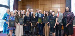 Group portrait of about 20 faculty and staff standing indoors at an awards ceremony, smiling toward the camera. Several people in the front row hold dark University of South Carolina award folders, with wood-paneled walls and large windows in the background.