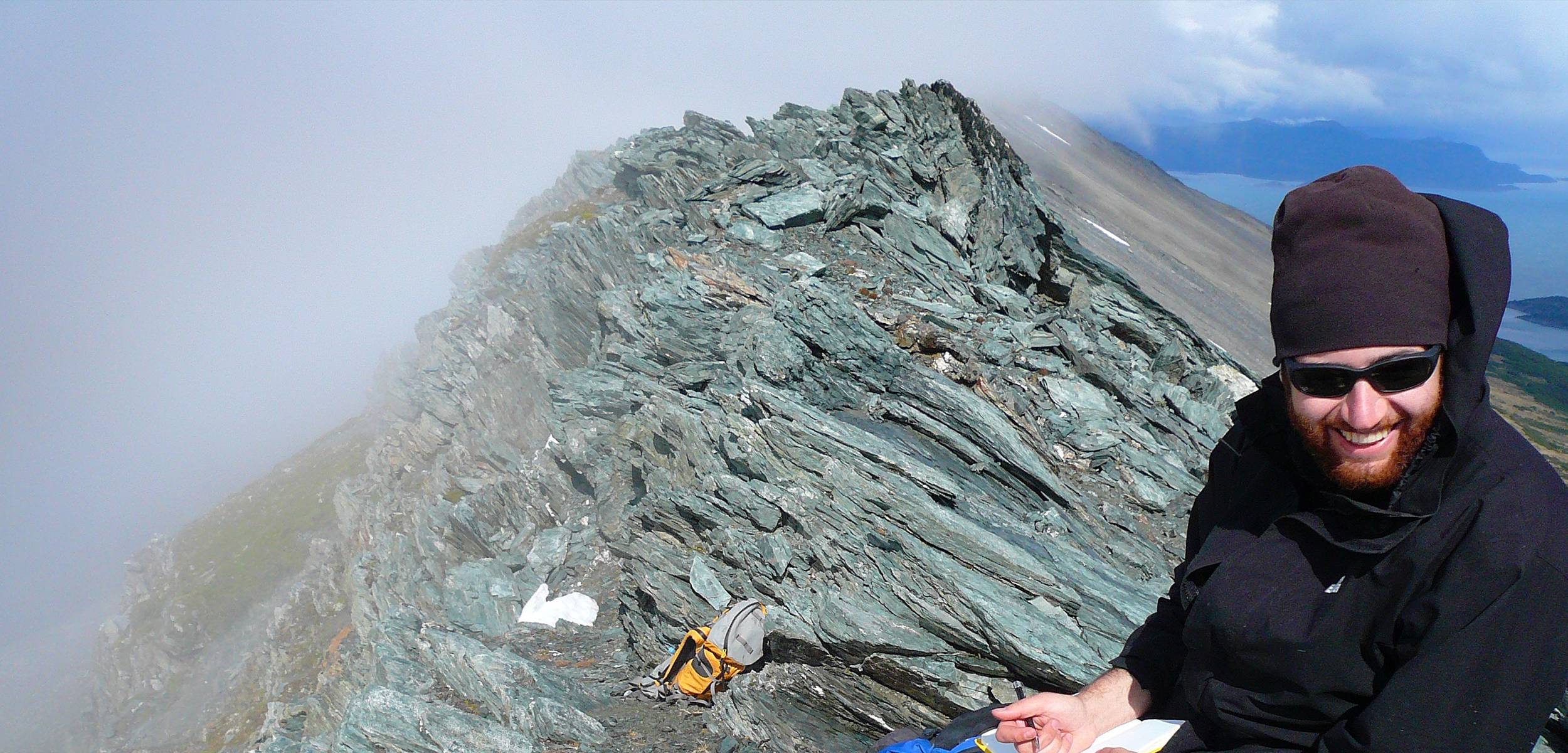 Person in dark hat and sunglasses smiling beside a rocky, windswept outcrop.