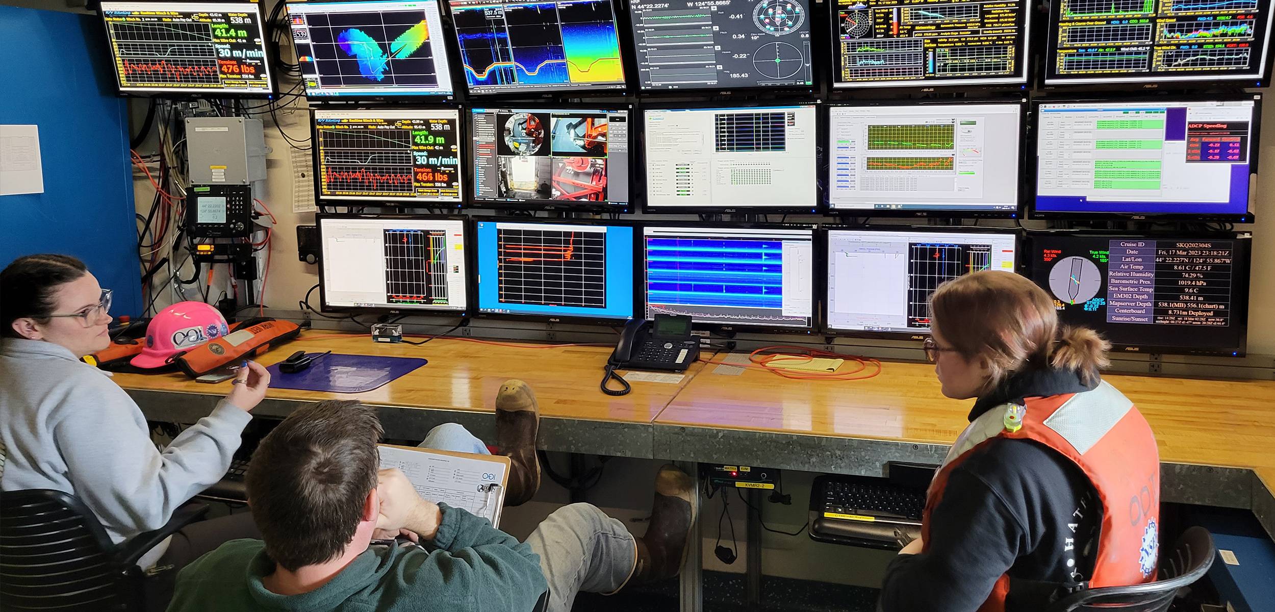 Control room on a research vessel with a wall of data monitors; three team members seated reviewing live readouts.