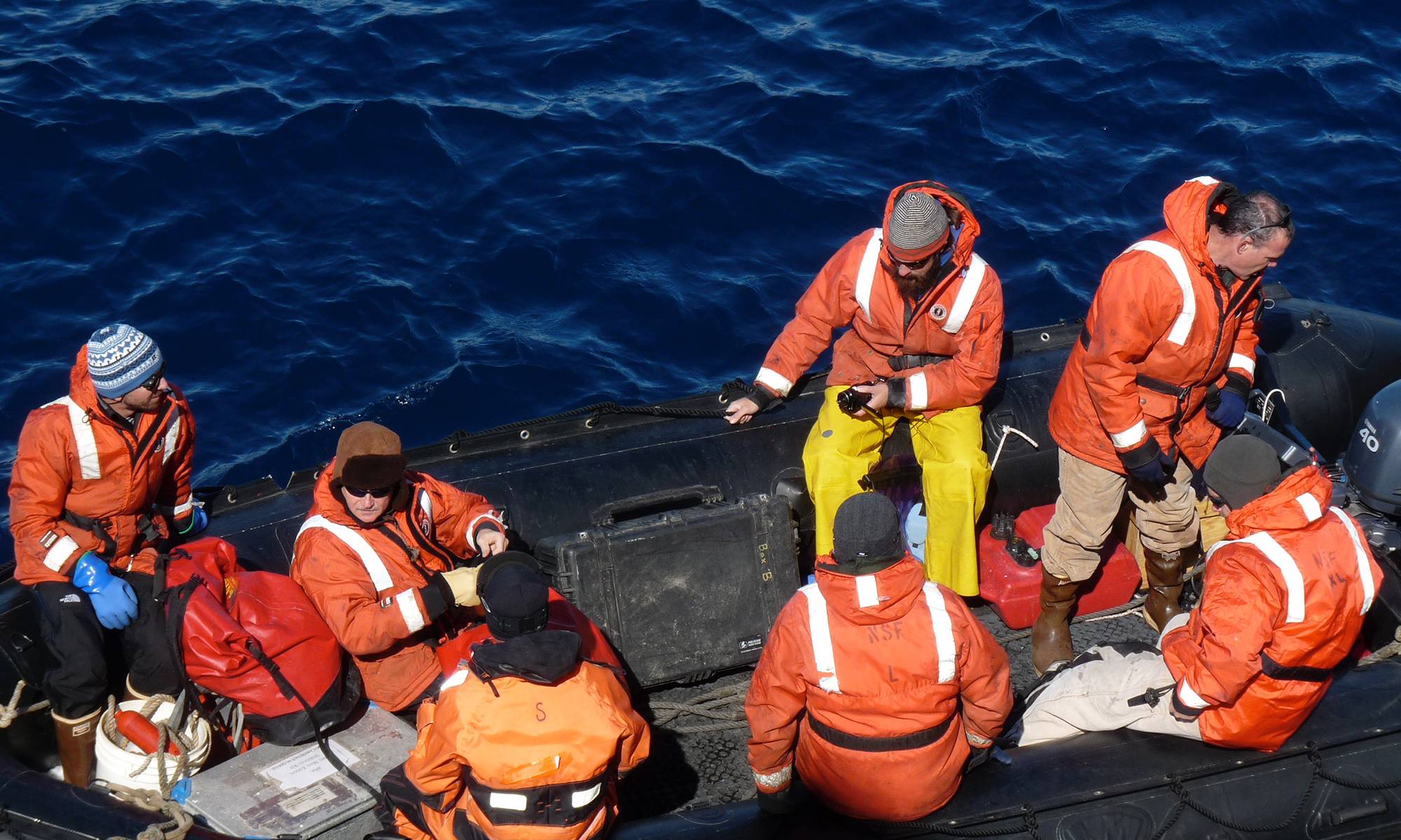 Research team in orange cold-weather gear on an inflatable boat over open ocean, preparing equipment.