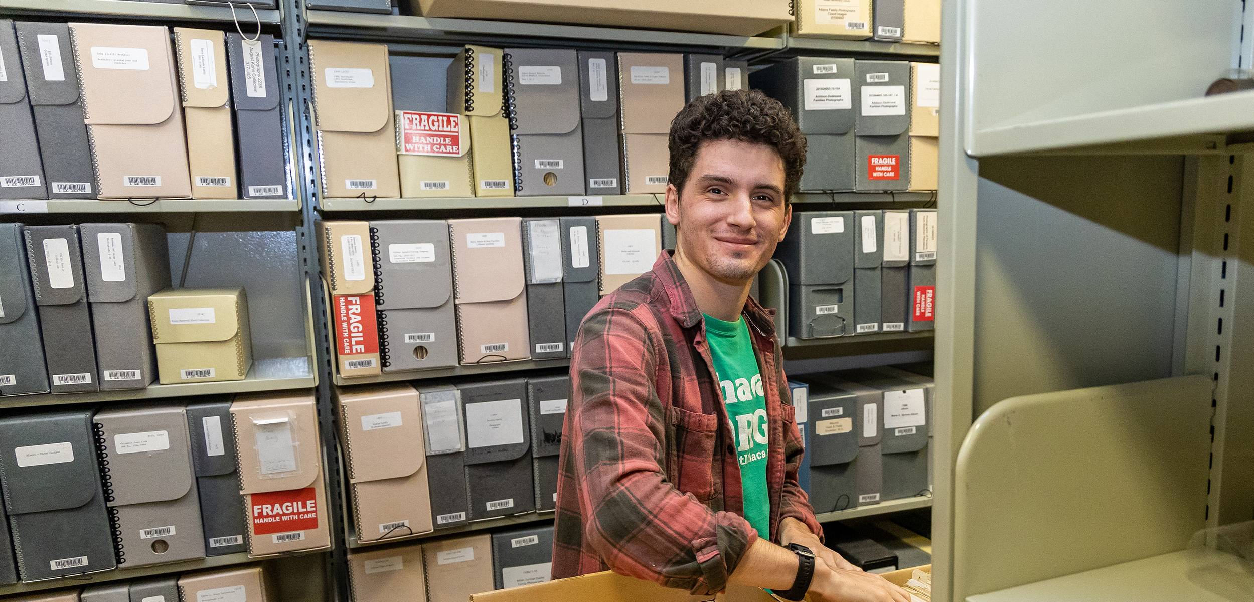 Student working in an archival storage room, standing between shelves of labeled document boxes and smiling at the camera.