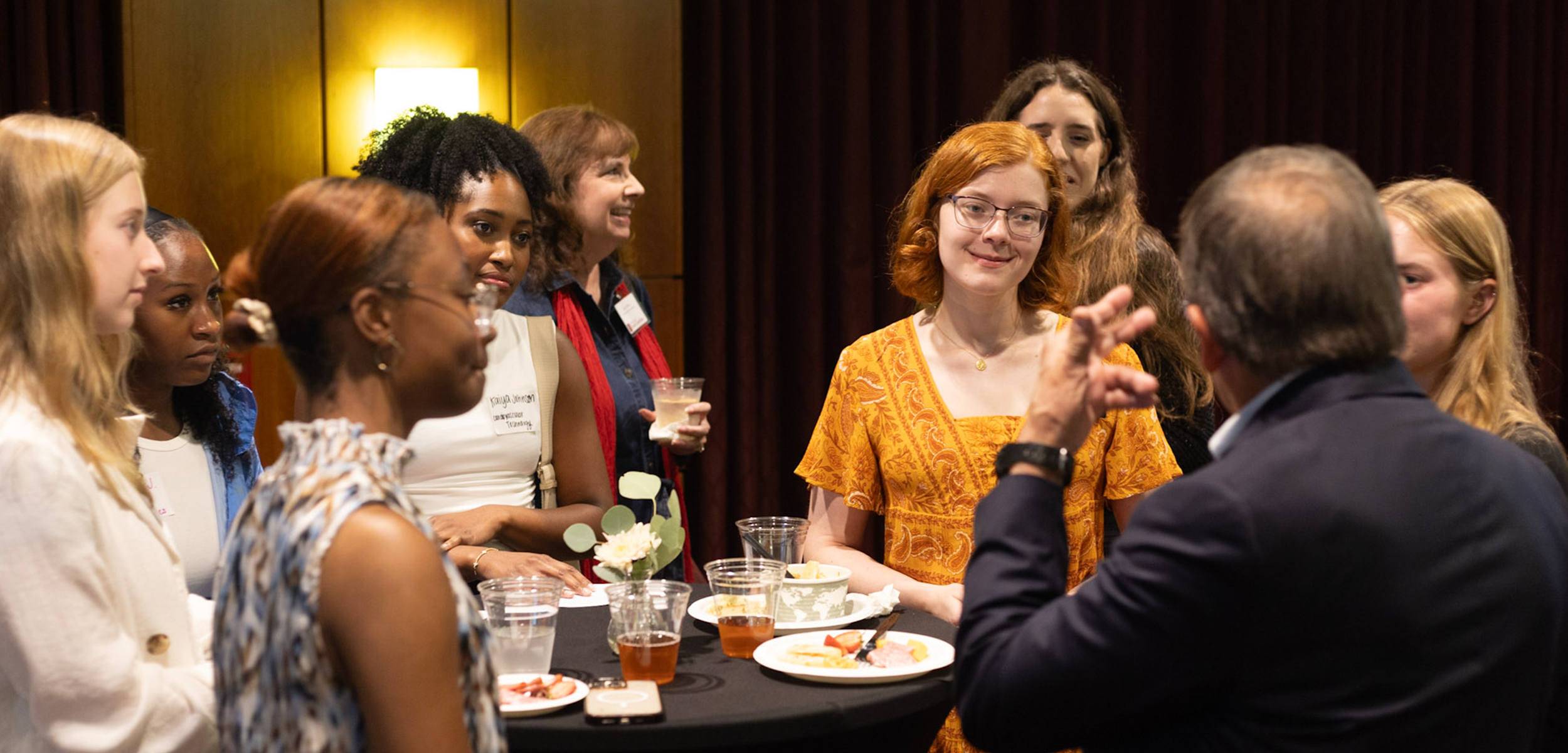 Group of people engaged in conversation at an indoor event, with a young woman in a yellow dress smiling and listening.