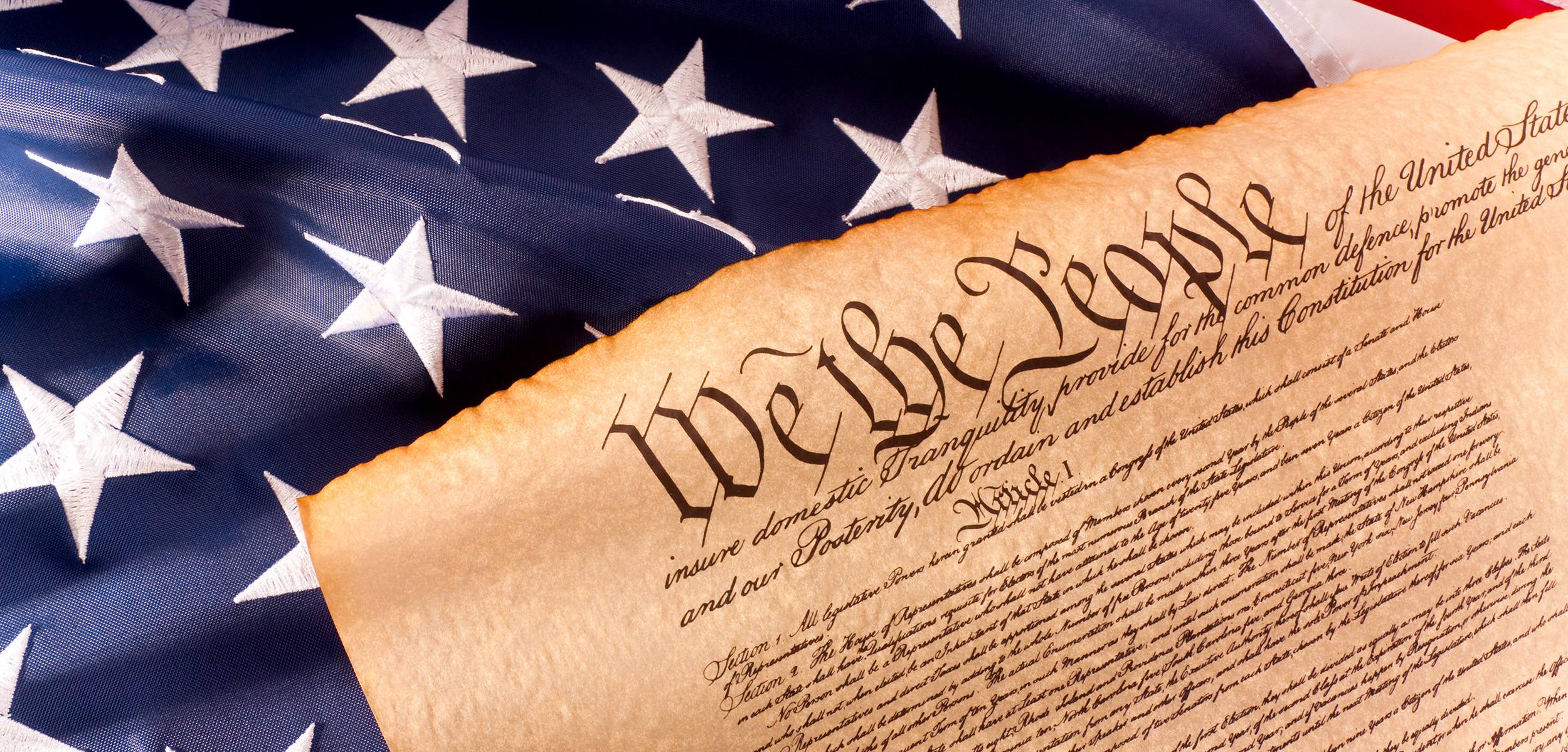 Close-up of the U.S. Constitution with the words 'We the People' visible, placed against a folded American flag with stars.