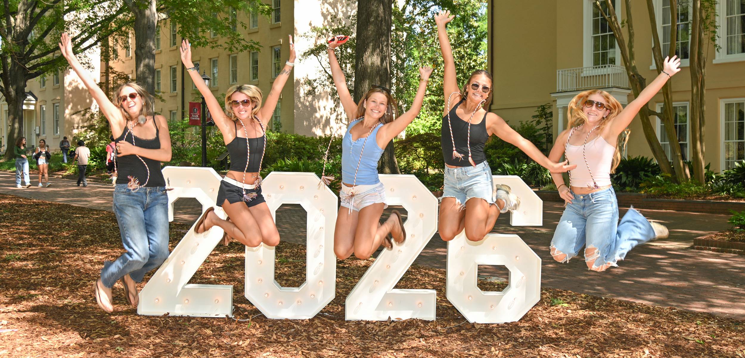 Five students jump behind large white 2026 numbers on a tree-lined campus lawn.