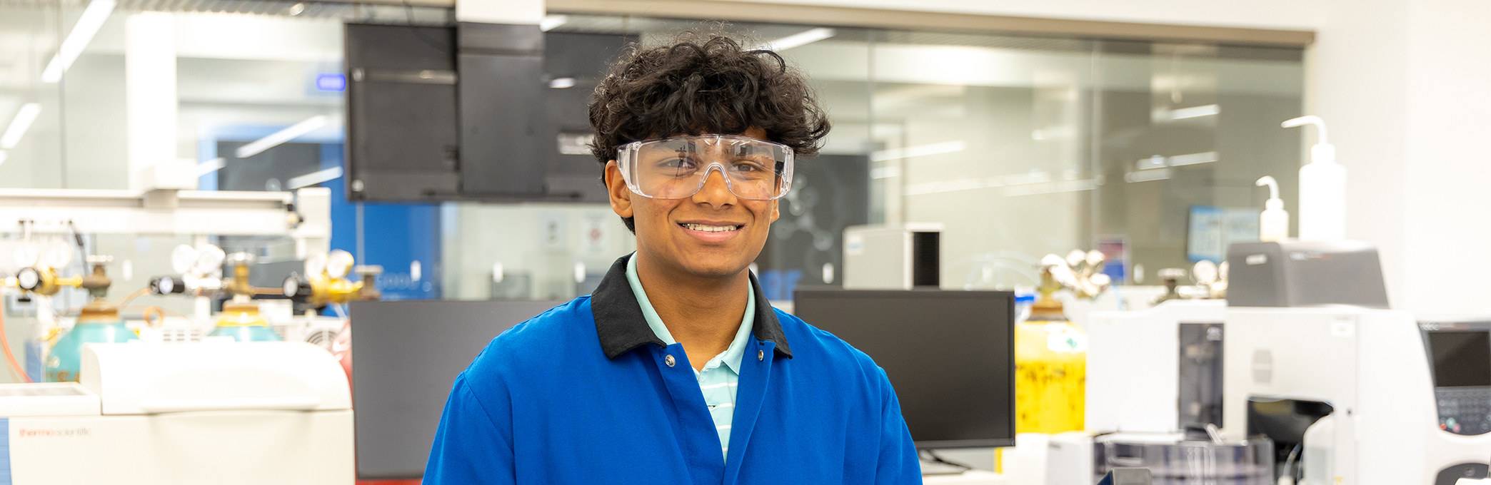Smiling student wearing safety goggles and a blue lab coat in a science laboratory.