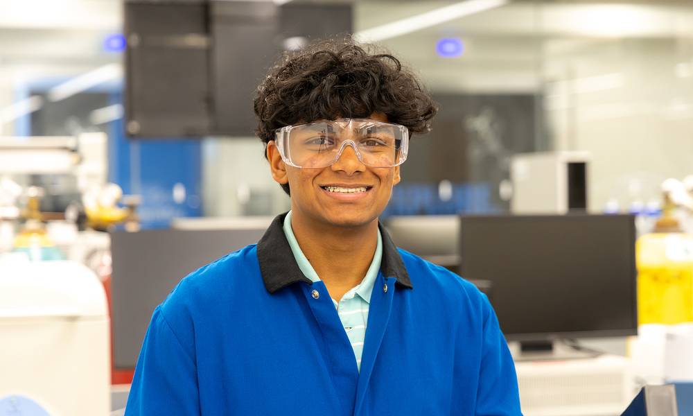 Smiling student wearing safety goggles and a blue lab coat in a science laboratory.