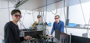 Three physics students wearing protective laser goggles work with cables and electronic equipment in a university optics lab.