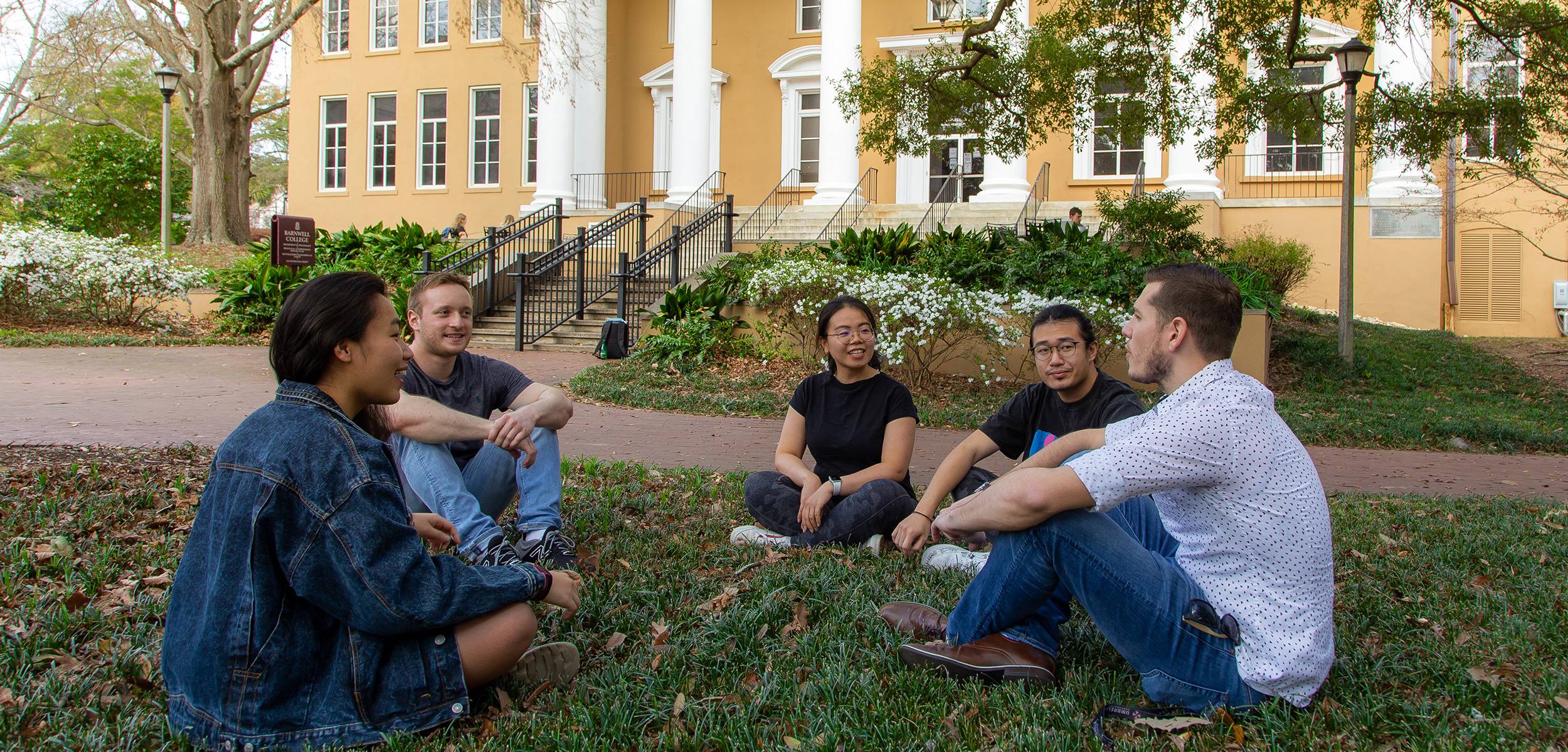 Five students sit together in a circle on a brick path in a campus garden, talking and smiling.