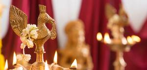 Close-up of an ornate brass bird-shaped ceremonial lamp wrapped with white flowers; candles and red drapery softly blurred behind.