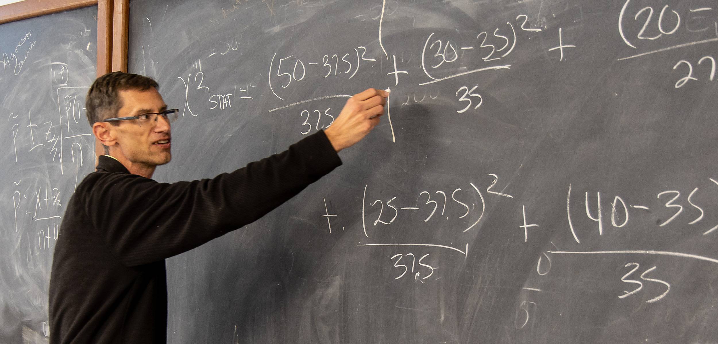 Instructor writing mathematical equations on a chalkboard during a statistics lecture.