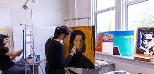 Art student paints a large portrait canvas in a bright studio with other vivid paintings nearby.