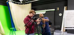 Instructor helps a student adjust a video camera on a tripod in a campus film studio.