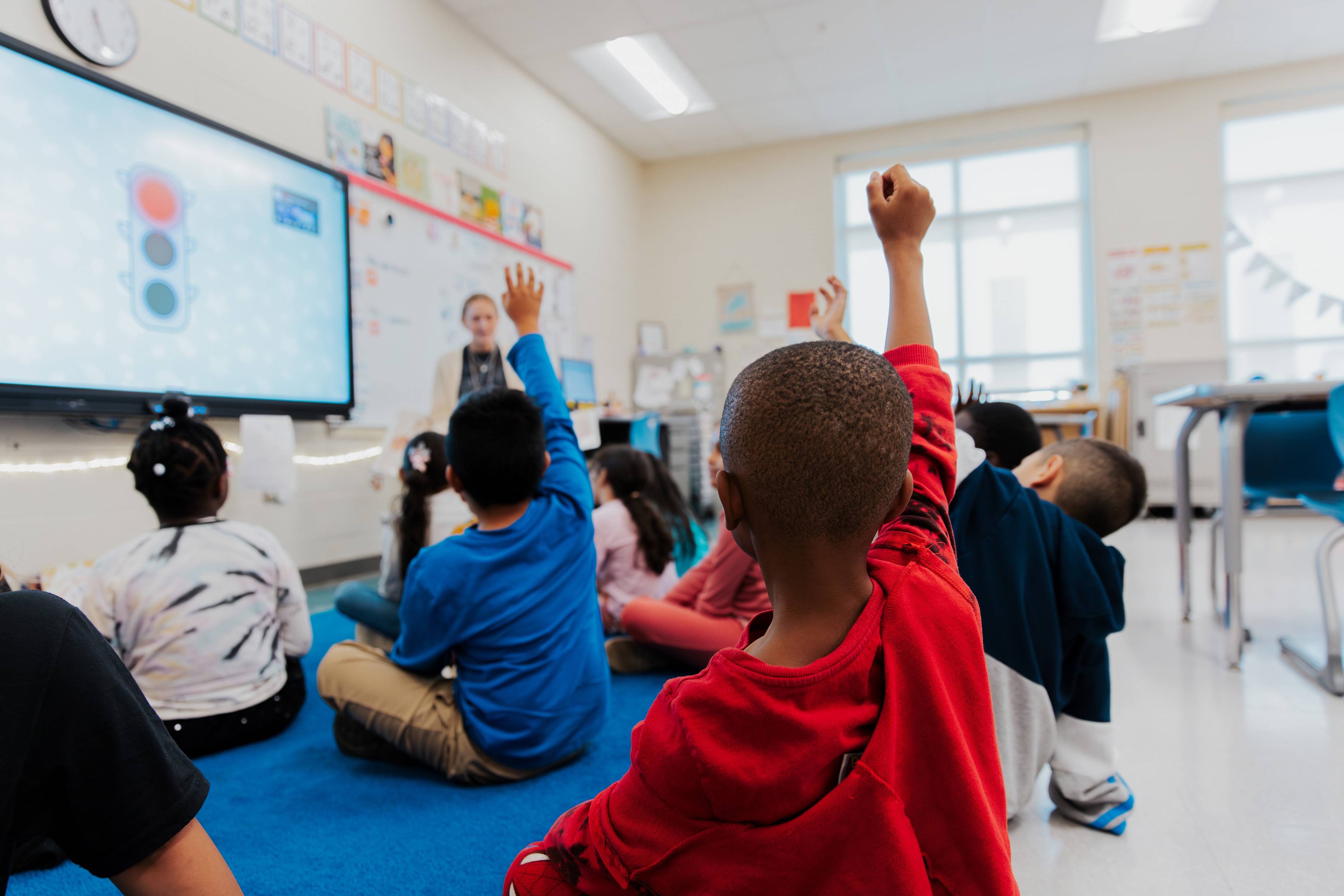 Elementary students in a classroom with hands raised.