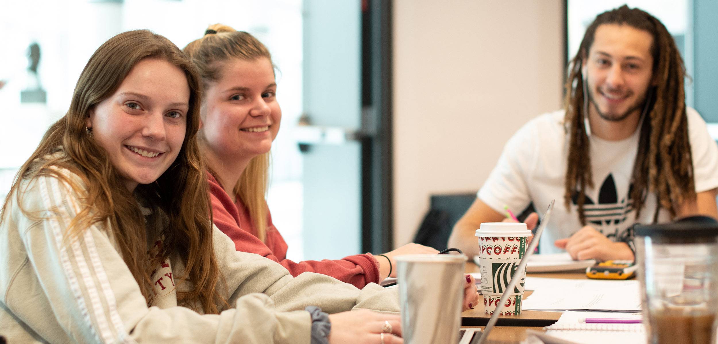 three students sitting at table with coffee studying, looking at camera