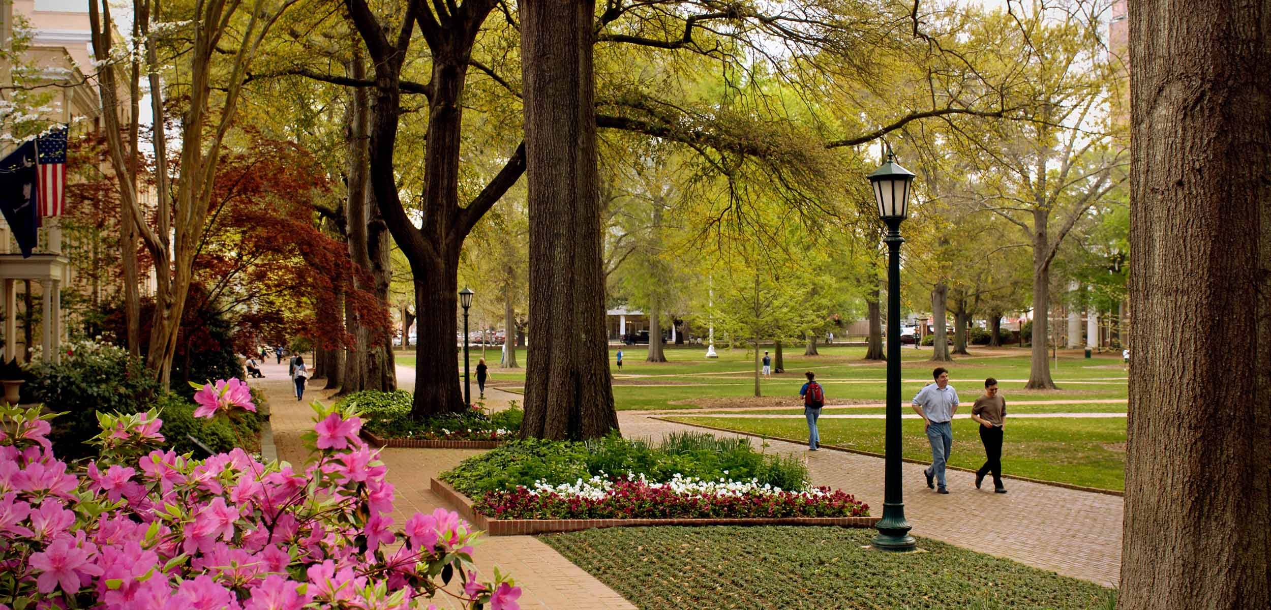 A scenic college campus walkway lined with large trees, blooming flowers, and American flags, as students stroll along brick paths on a sunny day.