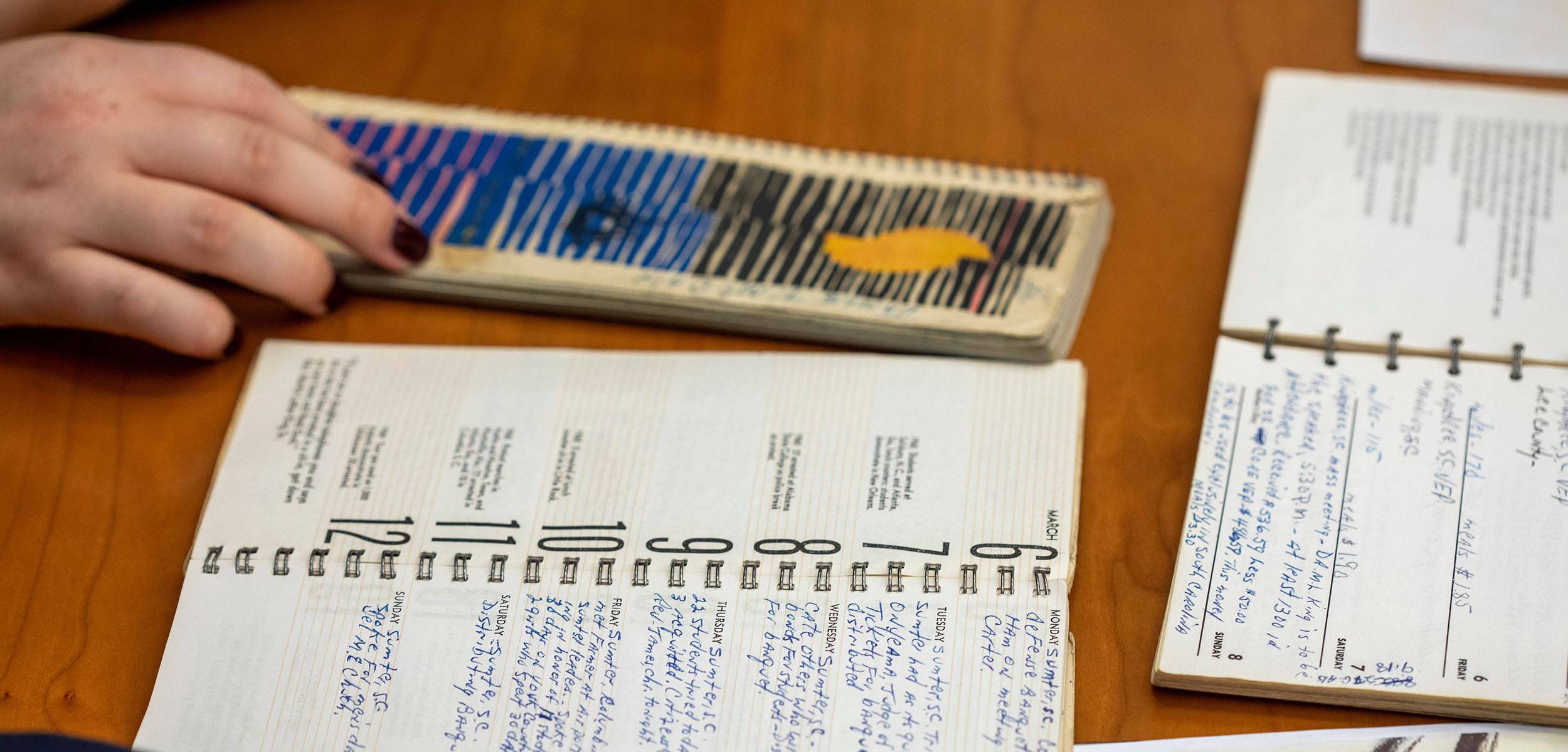 A hand rests on a colorful, striped notepad beside two open spiral-bound planners filled with handwritten notes, all placed on a wooden table.