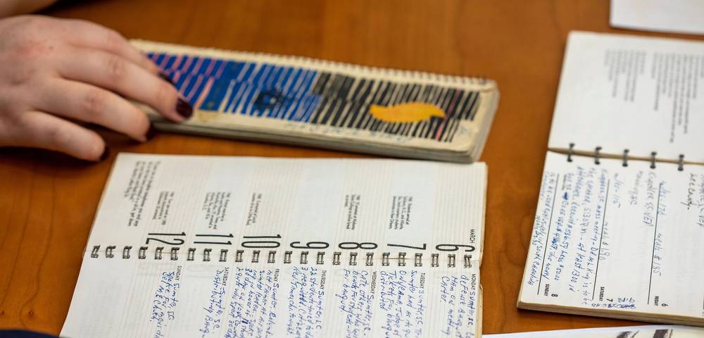 A hand rests on a colorful, striped notepad beside two open spiral-bound planners filled with handwritten notes, all placed on a wooden table.
