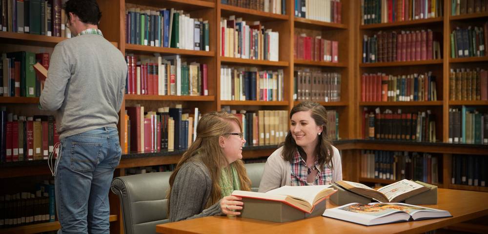 Two students sit at a library table smiling and studying large open books, while another person browses bookshelves in the background filled with colorful volumes.
