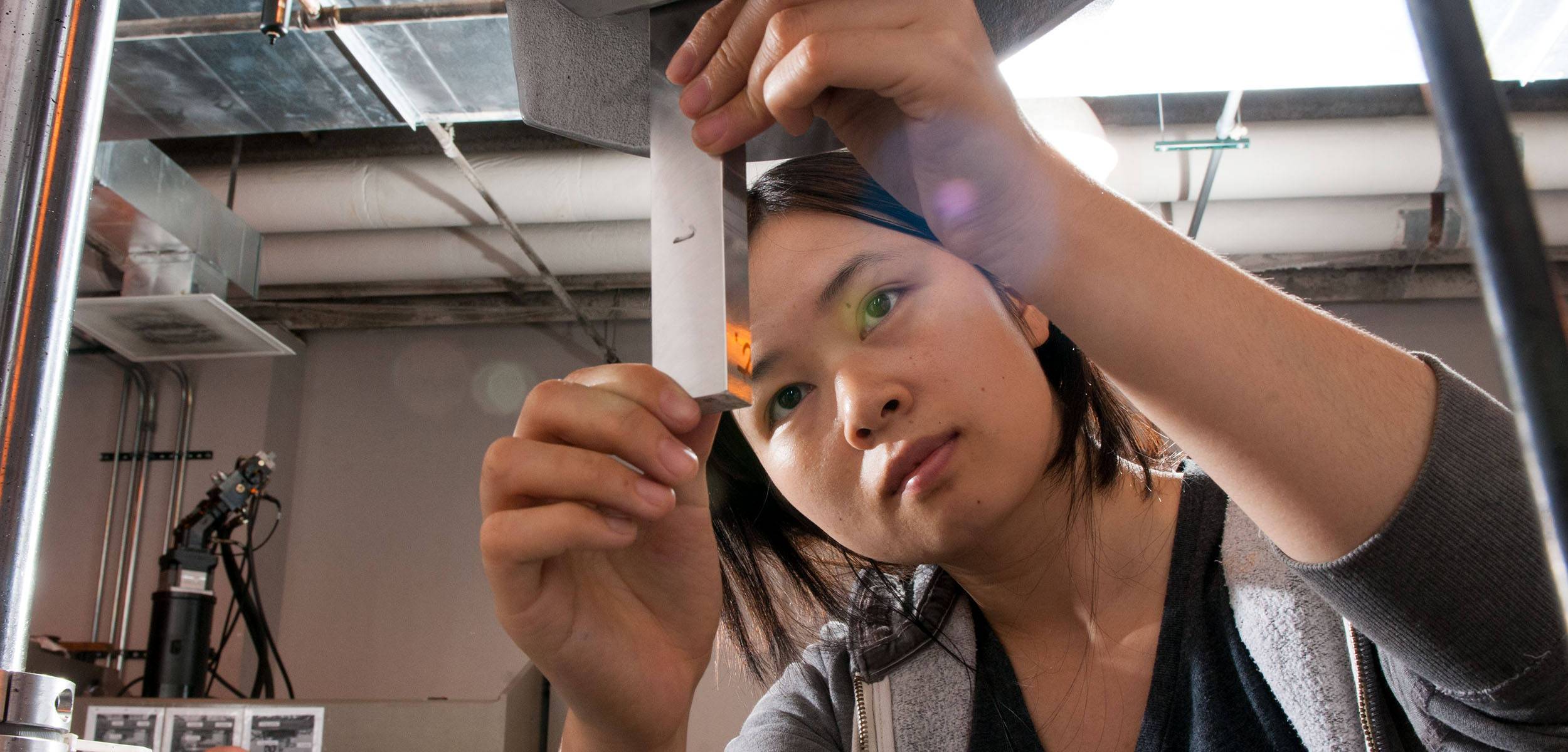 A focused student in a lab carefully adjusts a metallic apparatus, surrounded by industrial equipment and overhead pipes in a research environment.