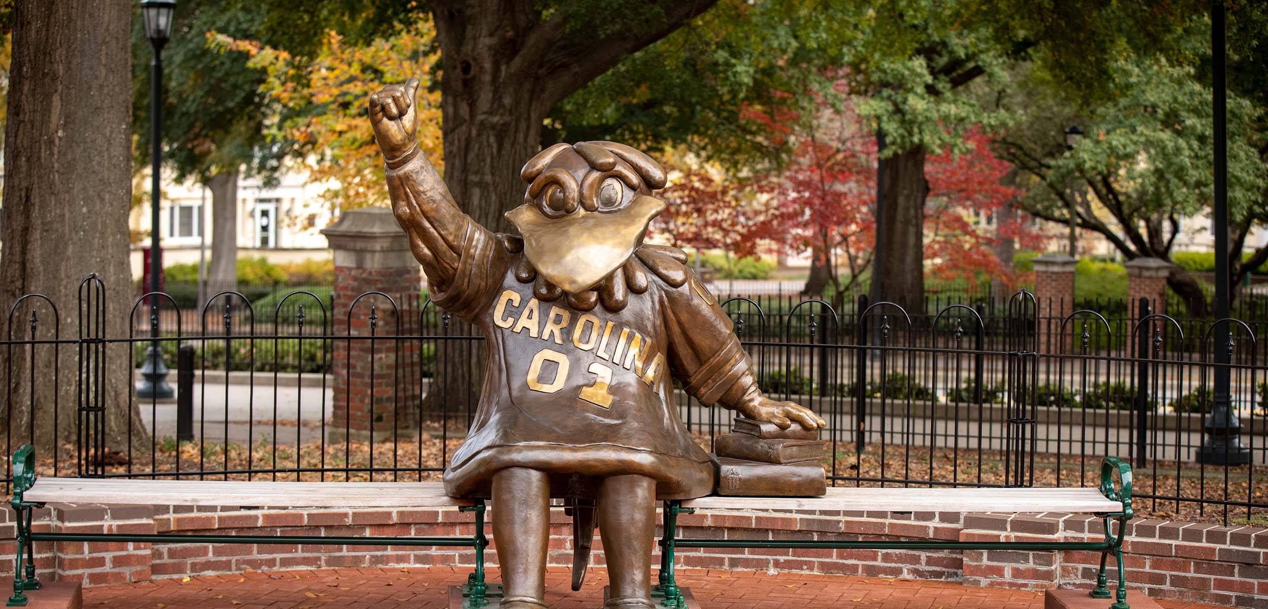 A bronze statue of the University of South Carolina mascot Cocky sits on a bench, wearing a jersey labeled 