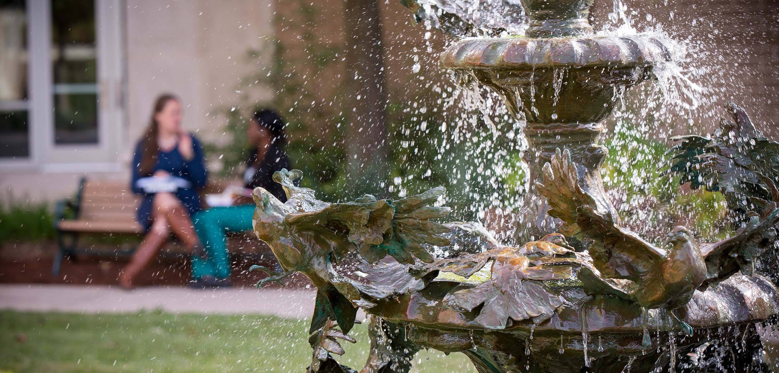 Close-up of a decorative bronze fountain with bird sculptures and flowing water, with two people talking on a bench in the blurred background.