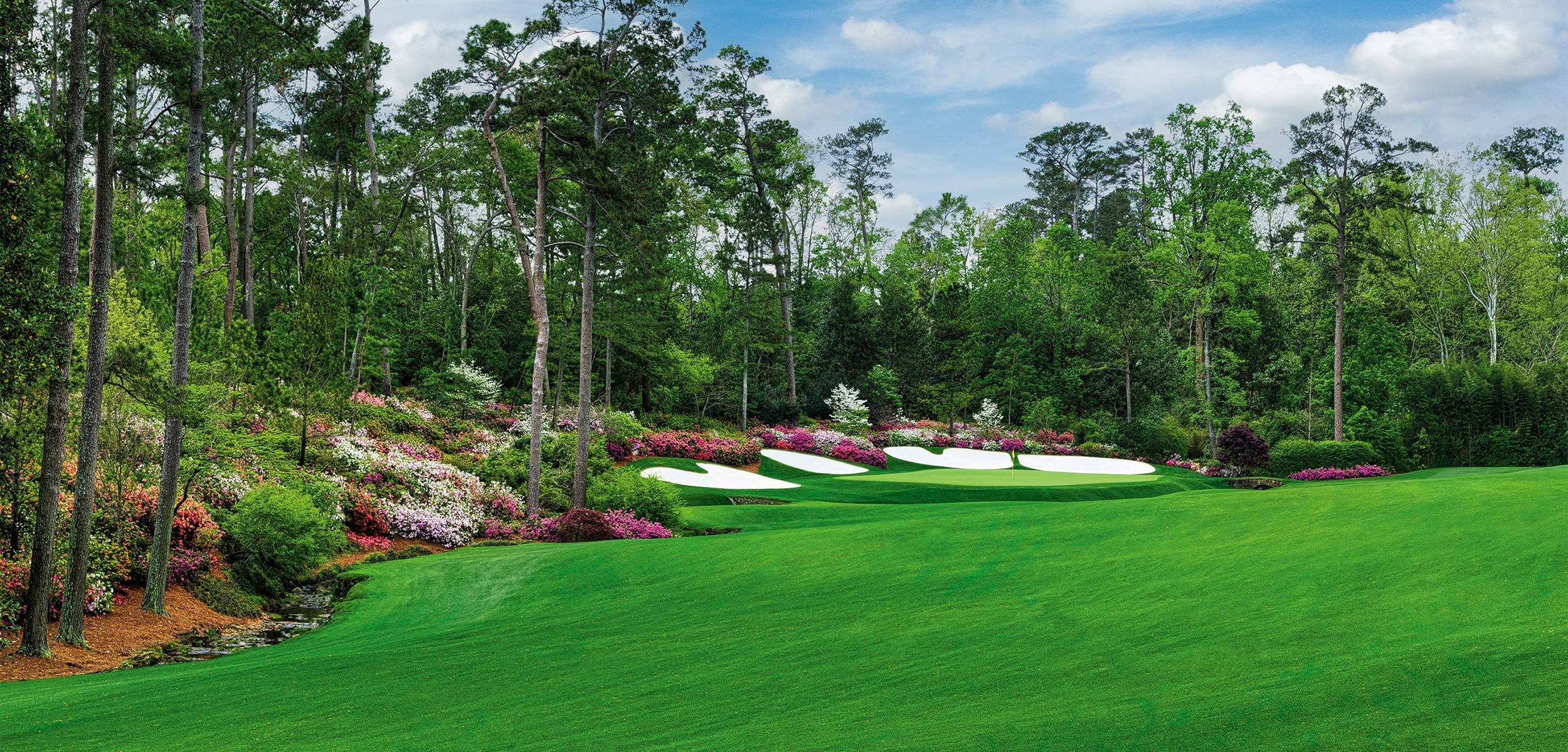 A lush green golf hole at Augusta National Golf Club bordered by towering pine trees, blooming pink and white azaleas, and bright white sand bunkers beneath a blue sky with scattered clouds. A small creek winds through the foreground, highlighting the course's meticulously maintained landscape.