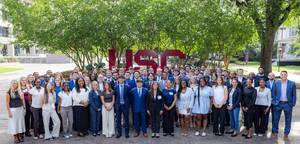 Group of sport and entertainment master's students dressed in business attire pose outdoors on the University of South Carolina campus in front of a large USC sign.