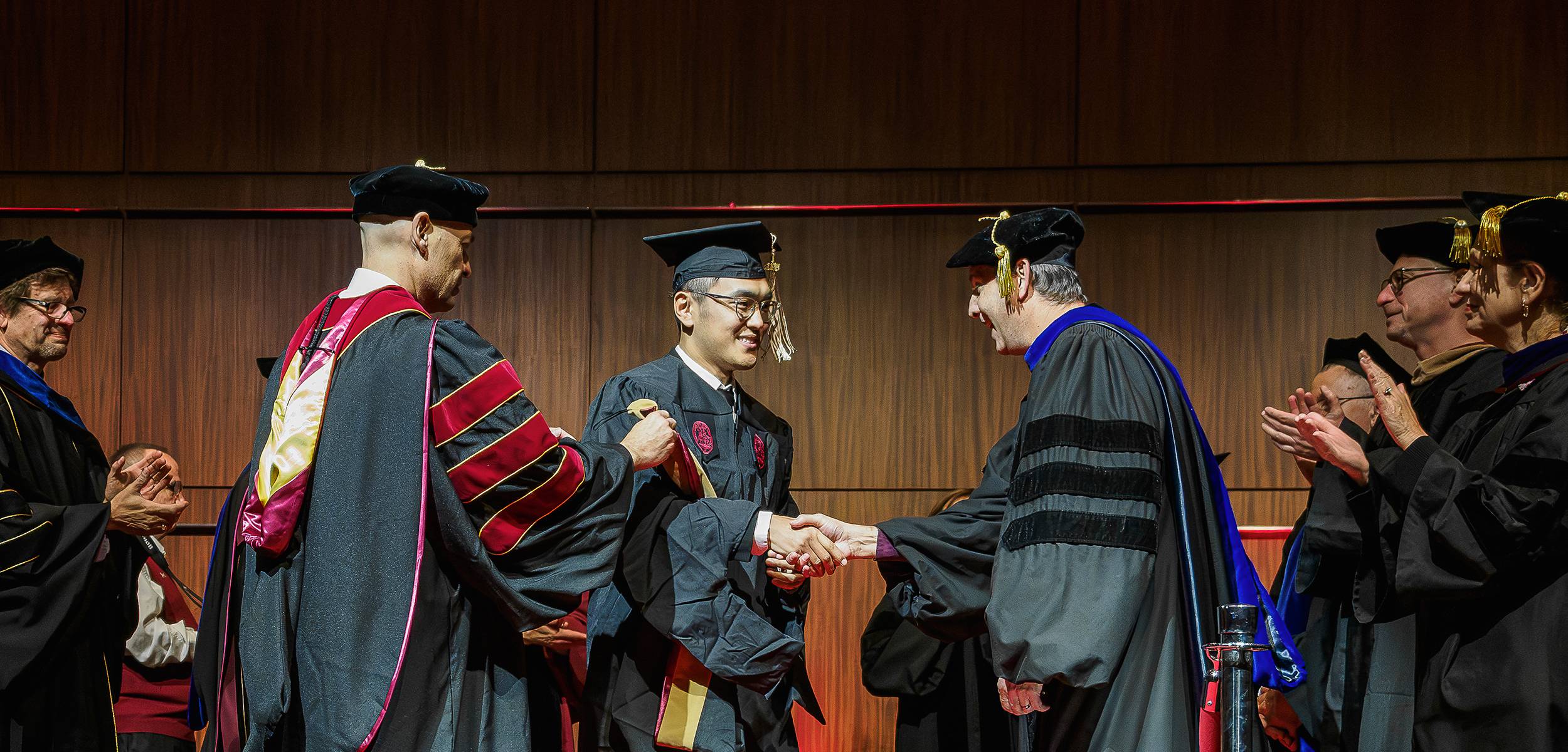 A graduate student in cap and gown shakes hands with a faculty member during a commencement ceremony as professors in academic regalia applaud on stage.