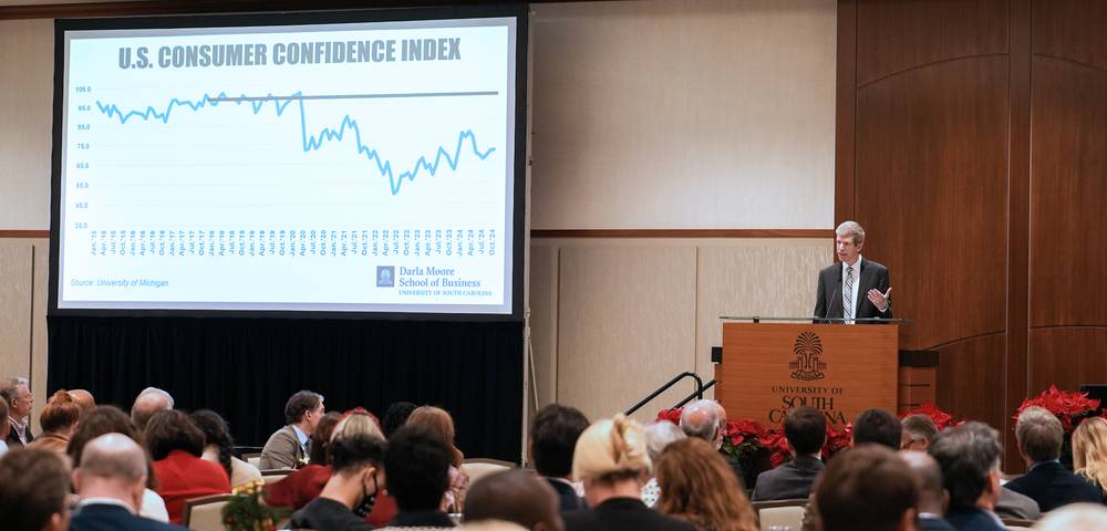 Moore School economist Joey Von Essen speaking at the annual Economic Outlook Conference; data on screen behind him.