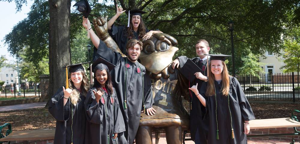 Group of USC students wearing graduations robes surrounding a statue of Cocky.