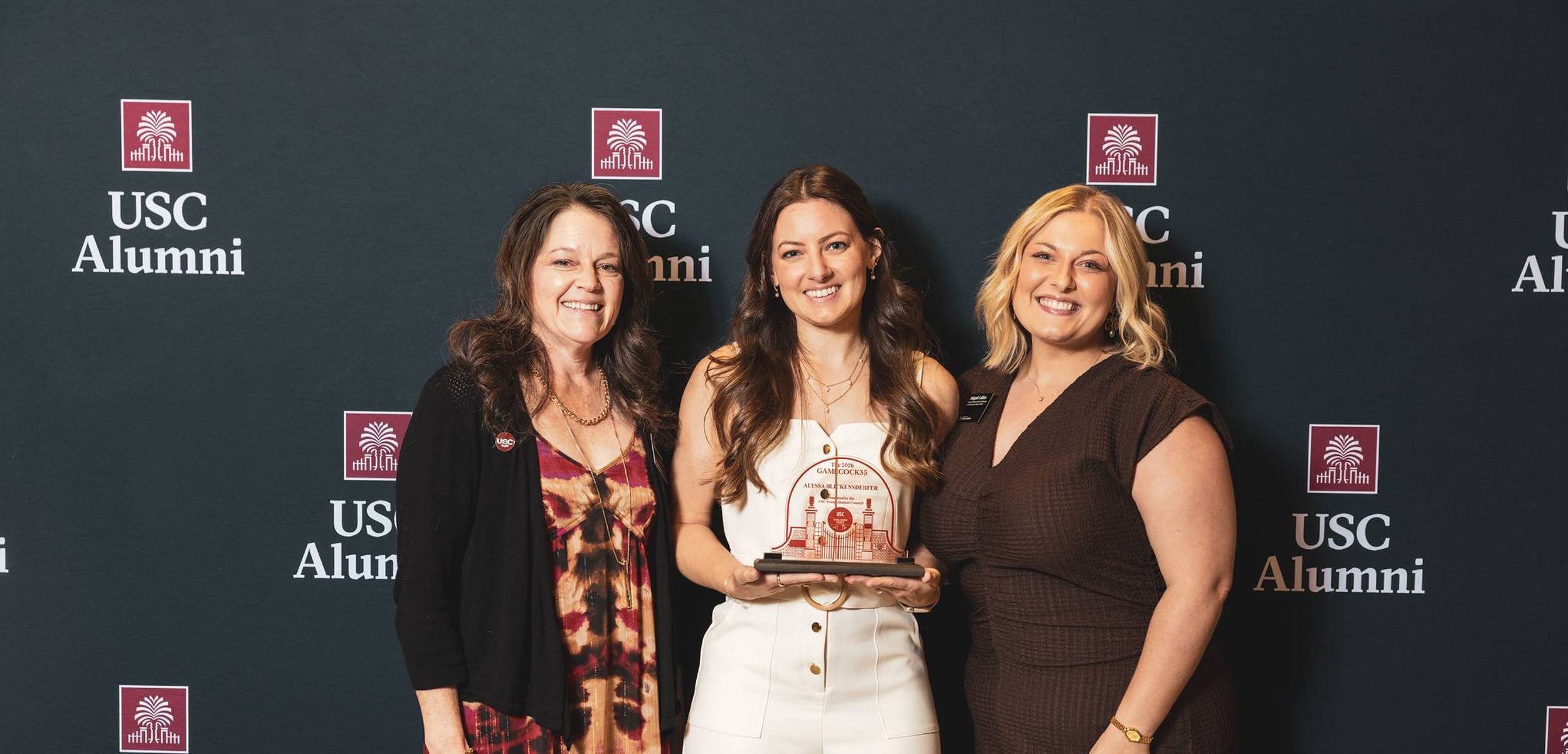Three women posing in front of a USC alumni banner